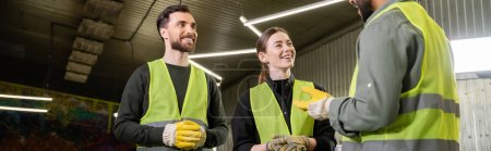 Photo for Cheerful workers in protective gloves and high visibility vests looking at indian colleague talking while standing together in blurred waste disposal station, recycling concept, banner - Royalty Free Image