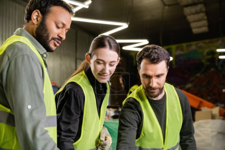 Photo for Cheerful workers in high visibility vests looking away while indian colleague talking and standing together in blurred garbage sorting center, garbage sorting and recycling concept - Royalty Free Image
