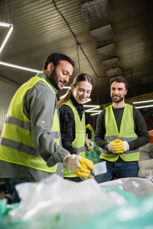 Joyeux jeune travailleur en gilet et gants haute visibilité debout près de collègues multiethniques près des sacs dans une station d'élimination des déchets floue, processus de tri des ordures 