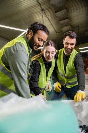 Lächelnde multiethnische Arbeiter in Schutzkleidung und Handschuhen reden und arbeiten in der Nähe verschwommener Säcke zusammen in der Müllentsorgungsstation, Mülltrennung 