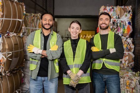 Photo for Cheerful multiethnic workers in high visibility vests and gloves crossing arms and looking at camera while standing near waste paper on disposal station, garbage sorting process - Royalty Free Image