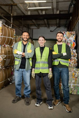 Smiling and interracial workers in high visibility vests and gloves crossing arms and looking at camera while standing near waste paper on disposal station, garbage sorting process 