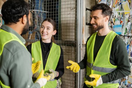 Cheerful man in high visibility vest and gloves talking to multiethnic colleagues while standing and resting in garbage sorting center, waste sorting and recycling concept