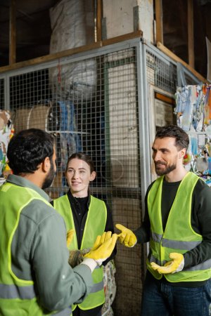 Photo for Smiling male worker in safety vest and gloves talking to indian colleague while resting near waste paper in garbage sorting center, waste sorting and recycling concept - Royalty Free Image
