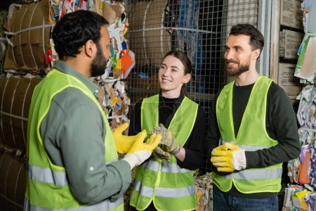 Photo for Indian worker in high visibility vest and gloves talking to smiling colleagues while standing near waste paper in blurred garbage sorting center, waste sorting and recycling concept - Royalty Free Image