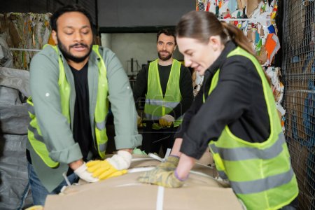 Smiling worker in reflective vest and gloves standing near hand pallet truck while blurred interracial colleagues pressing waste paper in garbage sorting center, waste sorting and recycling concept