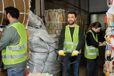 Smiling worker in protective vest and gloves standing near hand pallet truck and next to multiethnic colleagues working with waste paper in garbage sorting center, waste sorting and recycling concept