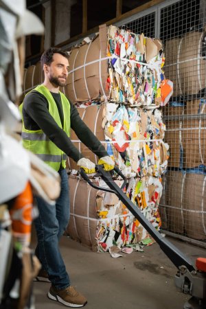 Bearded male worker in protective vest and gloves using hand pallet truck while standing near waste paper in garbage sorting center, waste sorting and recycling concept