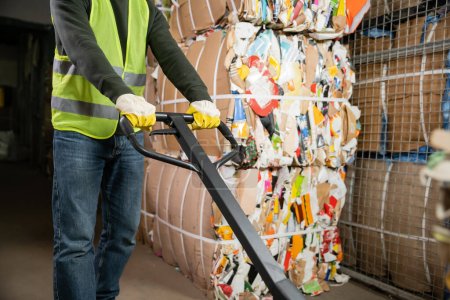 Photo for Cropped view of male sorter in protective gloves and vest using hand pallet truck while working near waste paper in blurred garbage sorting center, waste sorting and recycling concept - Royalty Free Image