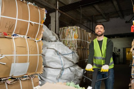 Travailleur souriant et barbu en gilet réfléchissant et gants à l'aide d'un transpalette à main tout en déplaçant les déchets de papier dans un centre de tri des déchets flou, concept de tri et de recyclage des déchets