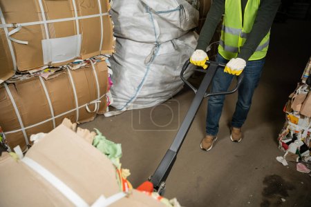 Photo for Cropped view of sorter in high visibility vest and gloves using hand pallet truck and moving waste paper in garbage sorting center, waste sorting and recycling concept - Royalty Free Image