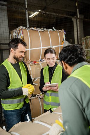 Cheerful young woman in safety vest using digital tablet near multiethnic colleagues while working with waste paper in garbage sorting center, waste sorting and recycling concept