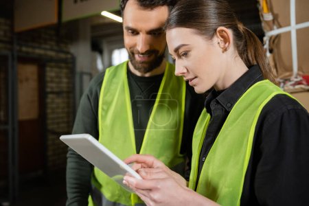Young worker in safety reflective vest using digital tablet while standing near smiling colleague and blurred waste paper in garbage sorting center, waste sorting and recycling concept