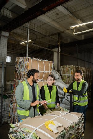 Positive indian worker in protective vest using digital tablet and talking to colleagues while standing near waste paper on hand pallet truck in waste disposal station, garbage sorting concept