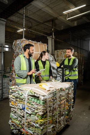 Cheerful sorter in protective vest and gloves talking to interracial colleagues with digital tablet and waste paper on hand pallet truck in waste disposal station, garbage recycling concept