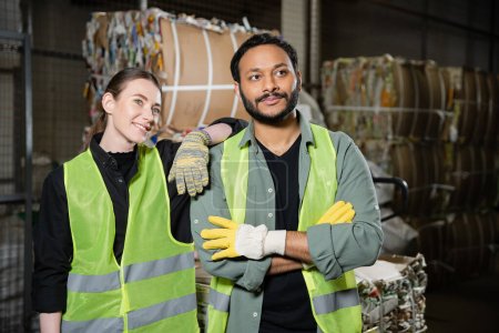 Cheerful young worker in protective vest and glove looking away while standing near indian colleague and blurred waste paper in waste disposal station, garbage sorting and recycling concept