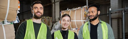 Cheerful and interracial workers in high visibility vests looking at camera while standing near waste paper in blurred waste disposal station, garbage sorting and recycling concept, banner 