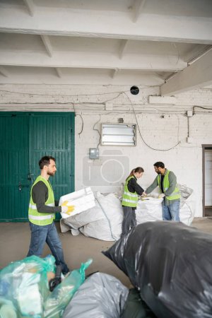 Male worker in high visibility vest and gloves carrying foam plastic while interracial colleagues working with sacks in waste disposal station, garbage sorting and recycling concept