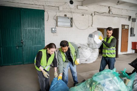 Foto de Trabajadores multiétnicos en chalecos y guantes de protección que trabajan con bolsas de plástico cerca de su colega en la estación de eliminación de residuos, clasificación de basura y el concepto de reciclaje - Imagen libre de derechos