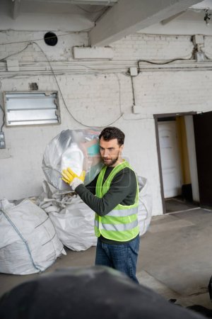 Trabajador barbudo en chaleco protector y guantes que transportan bolsa de plástico con basura mientras trabaja en la estación de eliminación de residuos, clasificación de basura y concepto de reciclaje