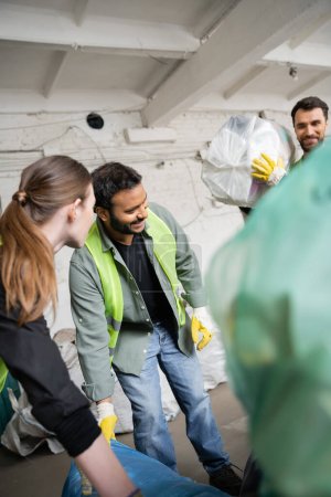 Trabajador indio sonriente en chaleco de alta visibilidad y guantes trabajando cerca de colegas y bolsas de plástico en la estación de eliminación de residuos, clasificación de basura y concepto de reciclaje