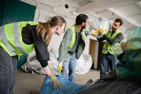 Trabajadora joven en chaleco protector y guantes sosteniendo bolsa de plástico cerca de colegas multiétnicos borrosos en la estación de eliminación de residuos, clasificación de basura y concepto de reciclaje