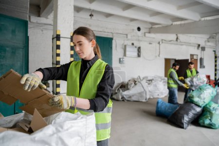 Junge Arbeiterinnen in Schutzweste und Handschuhen, die Pappe in der Nähe von Sack und Asche halten, und verschwommene Kollegen, die in der Müllentsorgungsstation, im Müllsortier- und Recyclingkonzept arbeiten
