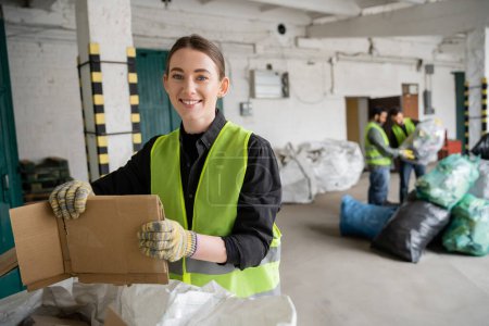 Wesoły młody pracownik w kamizelce ochronnej i rękawiczkach, trzymający tekturę i patrzący w kamerę podczas pracy w pobliżu worka i rozmytych kolegów w sortowni śmieci, koncepcja recyklingu