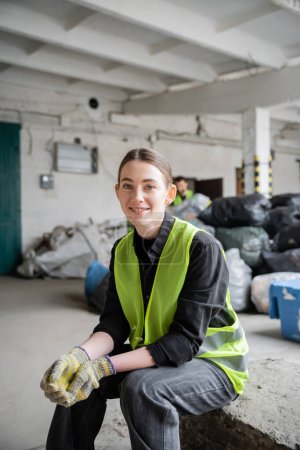 Positive young worker in protective vest and gloves looking at camera while resting near blurred plastic bags with trash at background in garbage sorting center, recycling concept