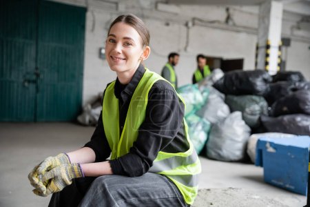 Wesoły młody pracownik w kamizelce bezpieczeństwa i rękawice patrząc w kamerę podczas odpoczynku i siedząc w pobliżu rozmazanych plastikowych toreb w centrum sortowania śmieci, koncepcja recyklingu