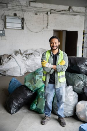 Foto de Trabajador indio alegre y barbudo en chaleco protector y guantes cruzando brazos y mirando a la cámara mientras está de pie cerca de bolsas de plástico en el centro de clasificación de basura, concepto de reciclaje - Imagen libre de derechos