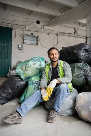 Téléchargez les photos : Travailleur indien masculin en gilet haute visibilité et gants de protection assis sur des sacs en plastique avec des déchets tout en travaillant dans le centre de tri des ordures, concept de recyclage - en image libre de droit