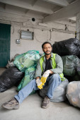 Male indian worker in high visibility vest and protective gloves sitting on plastic bags with trash while working in garbage sorting center, recycling concept puzzle #658270860
