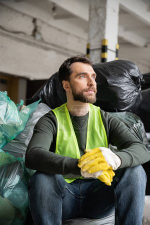 Bearded worker in high visibility vest and gloves looking away while sitting near plastic bags with trash in blurred garbage sorting center, recycling concept