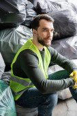 Bearded male worker in fluorescent vest and gloves looking away while sitting near blurred plastic bags with trash in garbage sorting center, recycling concept Stickers #658270902