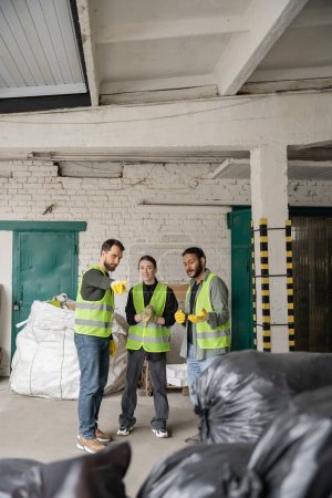 Bearded worker in protective vest and gloves pointing with finger at blurred plastic bags near smiling interracial colleagues in garbage sorting center, recycling concept