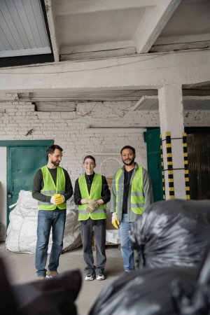 Positive multiethnic sorters in protective gloves and vests looking at camera while standing near blurred plastic bags with trash in garbage sorting center, recycling concept
