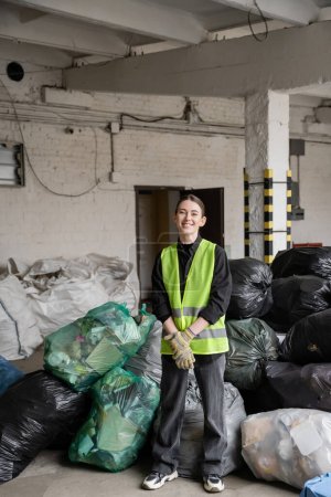 Trabajadora sonriente con guantes protectores y chaleco mirando a la cámara mientras está de pie cerca de bolsas de plástico con basura en el centro de clasificación de basura, concepto de reciclaje