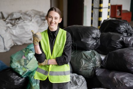 Lächelnder junger Arbeiter in Schutzweste und Handschuhen, der wie eine Geste wirkt und in die Kamera schaut, während er neben verschwommenen Plastiktüten mit Müll im Müllsortierzentrum steht, Recyclingkonzept