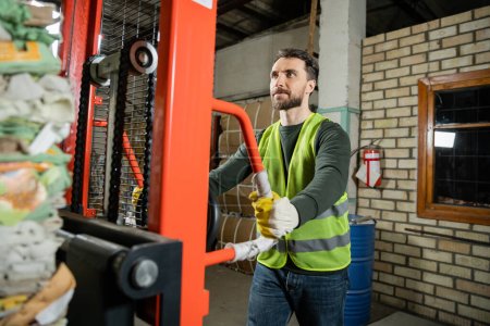 Photo for Bearded worker in fluorescent vest and gloves using hand pallet truck with waste paper while working in blurred garbage sorting center at background, recycling concept - Royalty Free Image