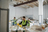 Male worker in protective vest and gloves holding plastic trash while standing near sacks and blurred interracial colleagues in waste disposal station, garbage sorting and recycling concept Longsleeve T-shirt #658271136