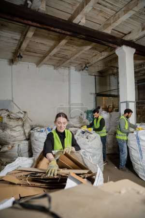 Trabajador positivo en guantes y chaleco poniendo cartón en el saco mientras separa la basura cerca de colegas multiétnicos borrosos en la estación de eliminación de residuos, clasificación de basura y concepto de reciclaje
