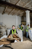 Positive worker in gloves and vest putting cardboard in sack while separating trash near blurred multiethnic colleagues in waste disposal station, garbage sorting and recycling concept Stickers #658271180
