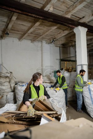 Young female worker in protective gloves and vest looking at multiethnic colleagues while putting cardboard in sack in waste disposal station, garbage sorting and recycling concept