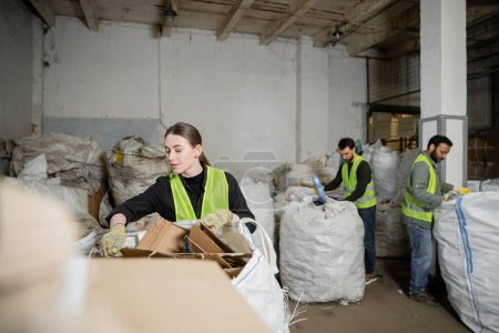 Young sorter in vest and protective gloves putting cardboard in sack near blurred multiethnic colleagues in waste disposal station, garbage sorting and recycling concept