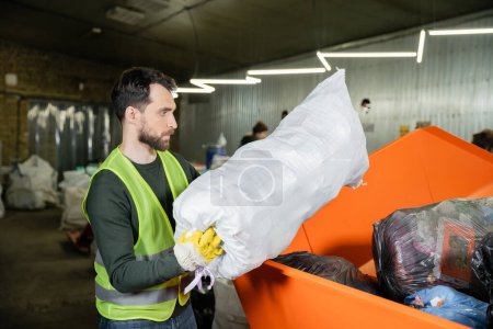 Photo for Side view of bearded sorter in glove and protective vest putting sack with trash in box in blurred waste disposal station, garbage sorting and recycling concept - Royalty Free Image