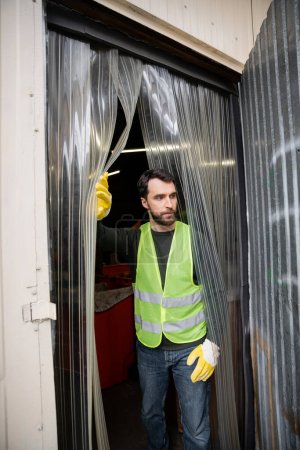 Bearded male worker in high visibility vest and gloves standing near entrance of waste disposal station, garbage sorting and recycling concept
