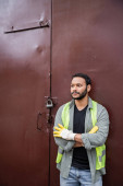 Bearded indian worker in high visibility vest and gloves crossing arms while standing near waste disposal station door, garbage sorting and recycling concept Stickers #658271398