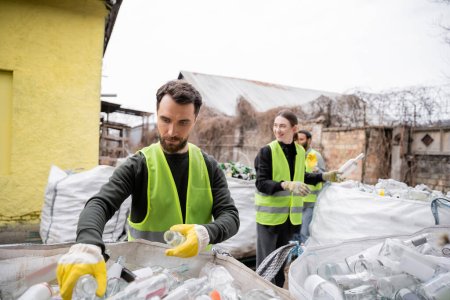 Bearded sorter in gloves and protective vest taking glass trash from sack near blurred and smiling multiethnic colleagues in outdoors waste disposal station, garbage sorting and recycling concept