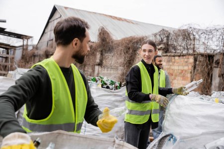 Trabajador sonriente en chaleco de seguridad y guantes sosteniendo basura de vidrio cerca de colegas multiétnicos y sacos en la estación de eliminación de residuos al aire libre, clasificación de basura y concepto de reciclaje